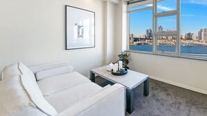 Ein modernes Wohnzimmer mit einer Couch, einem kleinen Tisch mit einer Pflanze und einem gerahmten Bild an der Wand. Ein großes Fenster bietet Blick auf eine Stadtlandschaft und ein Gewässer.