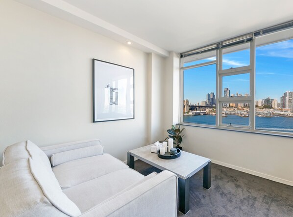 Un salon moderne avec un canapé, une petite table sur laquelle est posée une plante, et un tableau encadré au mur. Une grande fenêtre offre une vue sur les toits d’une ville et sur une étendue d’eau.