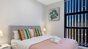 Une chambre moderne avec une tête de lit grise, un lit recouvert d’une couette et de draps blancs et roses, des oreillers à rayures et une vue sur la ville grâce à de grandes fenêtres.
