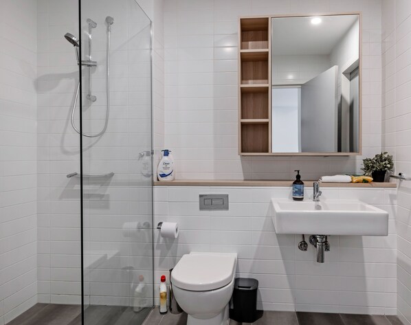 A modern bathroom with a glass shower enclosure, a white toilet, a sink with a mirror above it, and a small shelf with toiletries.
