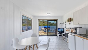 A compact kitchen with white cabinetry, a dining area with a round table and white chairs, and a view of a dock and water outside. - Kozyguru | Waterfront Holiday House With Kayak (Padstow Heights)