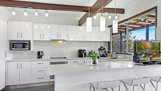 A modern kitchen with white cabinetry, a central island with bar stools, a coffee machine, and a large window with a view of trees. - Kozyguru | Waterfront Holiday House With Kayak (Padstow Heights)