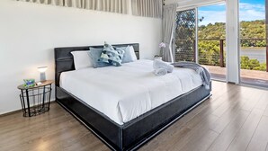 A spacious bedroom with a large bed, a ceiling fan, and a view of a lush landscape through the glass door. - Kozyguru | Waterfront Holiday House With Kayak (Padstow Heights)