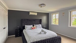 A bedroom with a large bed, a dark headboard, a bedside table, and a window with a view of greenery. - Kozyguru | Prestons Dream Holiday House + Parking (Prestons)