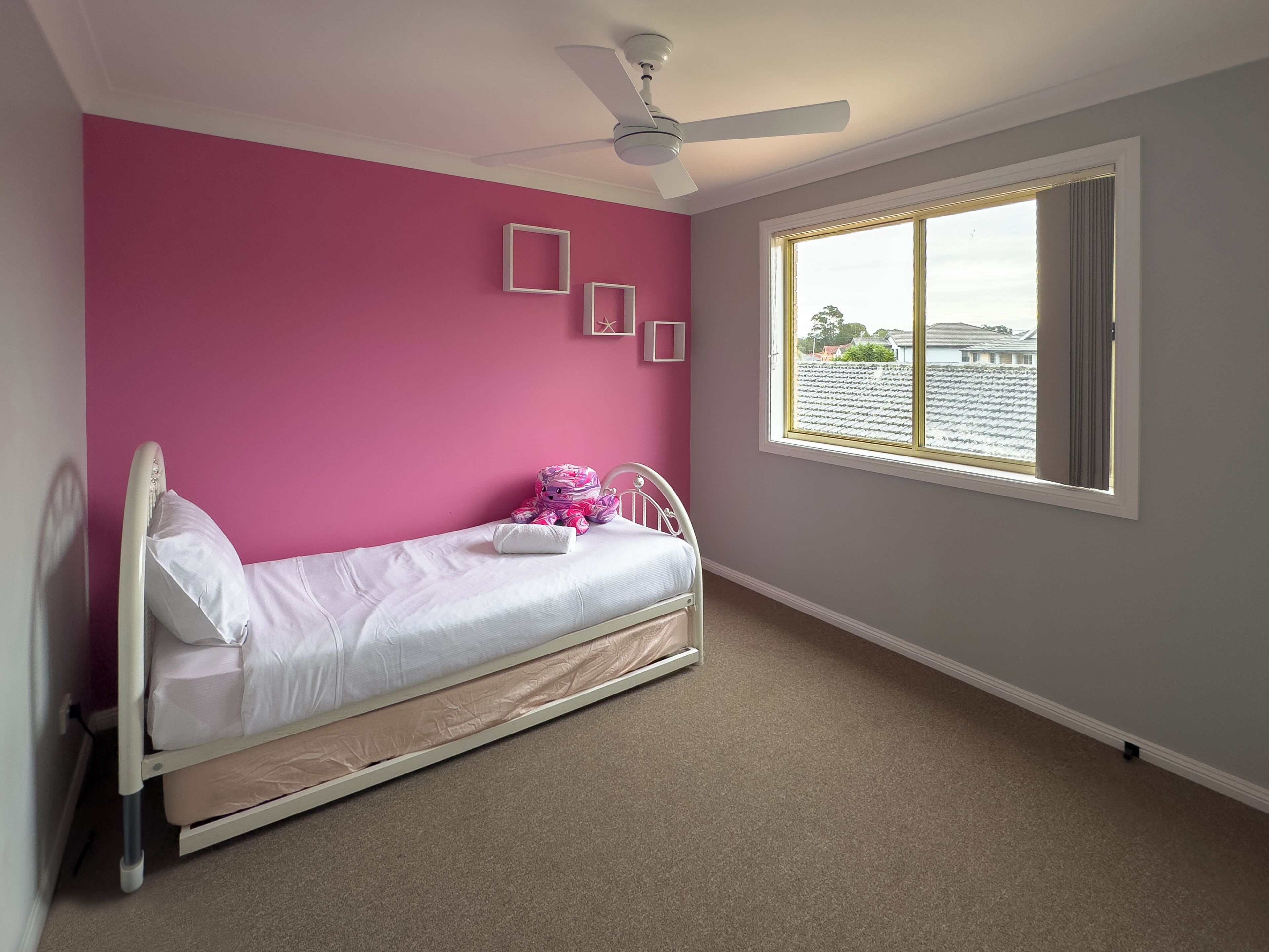 Un dormitorio con una cama individual, ventilador de techo, ventana con vista a zona residencial y pared rosa.