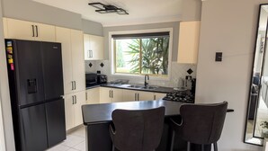 A modern kitchen with a black refrigerator, white cabinets, a gas stove, and two bar stools. - Kozyguru | Prestons Dream Holiday House + Parking (Prestons)