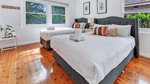 A bedroom with two beds, a white wall, a window, and a plant. - Kozyguru | Rockdale | Spacious 2 Bedrooms House (Rockdale)