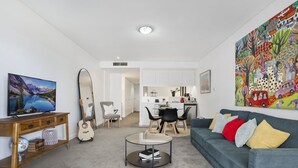 A modern living room with a sofa, coffee table, and a TV. There is a dining area with chairs and a kitchen in the background. - Kozyguru | Barangaroo | Waterfront Views Studio (Sydney)