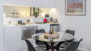 A modern kitchen with white cabinetry, a glass dining table, and a red kettle. - Kozyguru | Barangaroo | Waterfront Views Studio (Sydney)