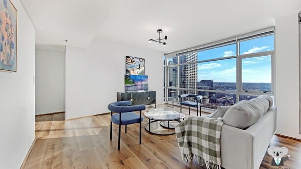 A modern living room with a grey sofa, blue armchairs, and a coffee table. A large window offers a city view. - Kozyguru | Sydneycbd | Amazing City View 3B Apt (Sydney)