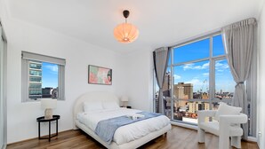 A modern bedroom with a large window, a bed, a chair, and a nightstand. - Kozyguru | Sydneycbd | Amazing City View 3B Apt (Sydney)