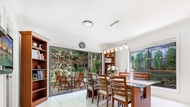 A dining area with a glass table and chairs, a wooden bookshelf, and a view of a garden through large windows. - Kozyguru | Westleigh | 4 Bed Room | Free Parking (Westleigh)