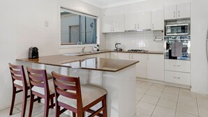 A modern kitchen with white cabinets, a large island with bar stools, a built-in oven, and a window above the sink. - Kozyguru | Westleigh | 4 Bed Room | Free Parking (Westleigh)