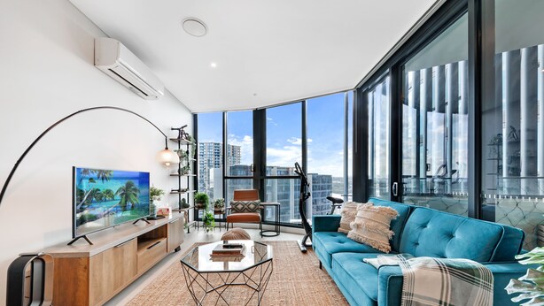 A modern living room with a large TV, a blue sofa, a coffee table, and a view of the city through large windows.