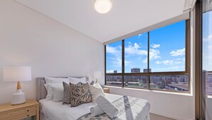 A bedroom with a large window, a bed with multiple pillows, a bedside table with a lamp, and a view of the cityscape. - Kozyguru | Wentworth Point | 2B Apt+Free Parking (Wentworth Point)