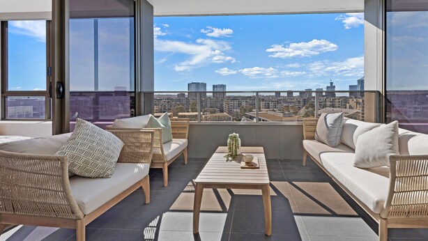 A modern balcony with wicker furniture, a wooden table, and a city view. - Kozyguru | Wentworth Point | 2B Apt+Free Parking (Wentworth Point)
