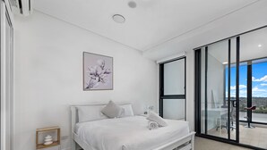 A modern bedroom with a white bed, a small bedside table, a wall-mounted artwork, and a large sliding glass door leading to a balcony with a view. - Kozyguru | Wentworth Point | 1 Bed Waterview Apt (Wentworth Point)