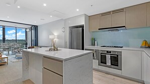 A modern kitchen with a central island, stainless steel appliances, and a view to the outdoors. - Kozyguru | Wentworth Point | 1 Bed Waterview Apt (Wentworth Point)