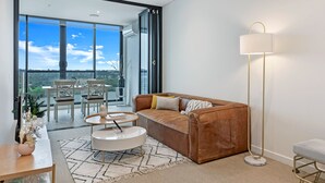A modern living room with a brown leather sofa, a white round coffee table, and a floor lamp. There is a dining area with a table and chairs, and a large window offering a view of the outdoors. - Kozyguru | Wentworth Point | 1 Bed Waterview Apt (Wentworth Point)