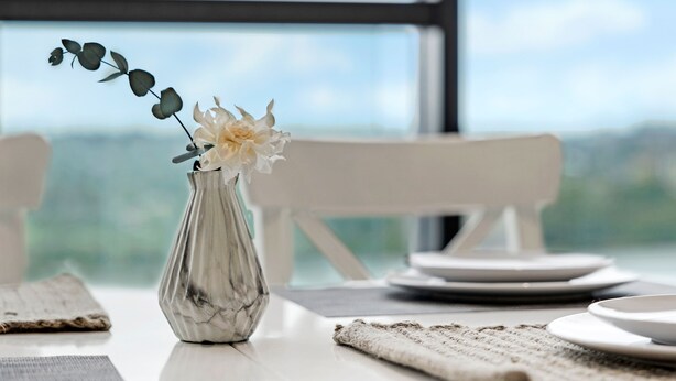 A table with a vase of flowers, a plate, and a view of the outdoors through a window. - Kozyguru | Wentworth Point | 1 Bed Waterview Apt (Wentworth Point)