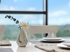 A table with a vase of flowers, a plate, and a view of the outdoors through a window. - Kozyguru | Wentworth Point | 1 Bed Waterview Apt (Wentworth Point)