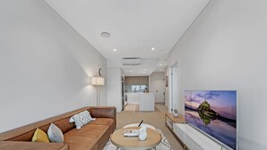 A modern living room with a brown leather sofa, a round coffee table, and a flat-screen TV displaying a scenic view. - Kozyguru | Wentworth Point | 1 Bed Waterview Apt (Wentworth Point)