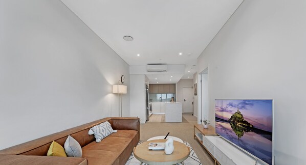 A modern living room with a brown leather sofa, a round coffee table, and a flat-screen TV displaying a scenic view. - Kozyguru | Wentworth Point | 1 Bed Waterview Apt (Wentworth Point)