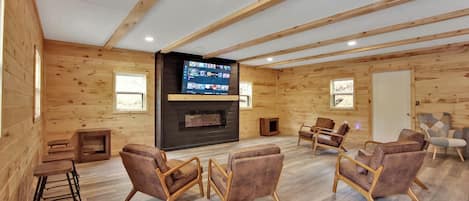 A spacious room with wooden walls and a large flat-screen TV mounted on a dark wall. There are several leather chairs arranged around a central area, and a small wooden table with stools.