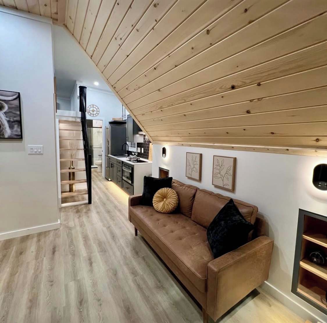 A modern living room with a brown leather sofa, wooden ceiling, and a kitchen in the background.