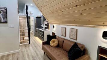 A modern living room with a brown leather sofa, wooden ceiling, and a kitchen in the background.