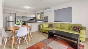 A modern living room with a green sofa, a dining table with chairs, a coffee table, and a kitchen in the background. - Kozyguru | Mt Gravatt East | 3 Bed Home (Mount Gravatt East)