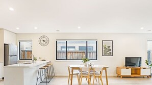 A modern kitchen with a central island, a dining area with a round table and chairs, a TV on a wooden cabinet, and a large window with a view of a building. - Kozyguru | Greenvale | Charming 7 Bed House (Greenvale)