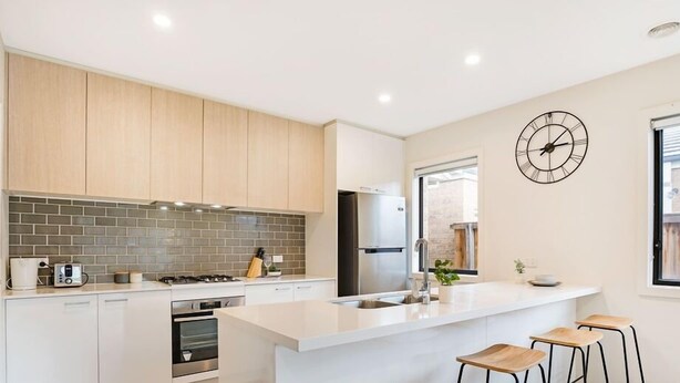 A modern kitchen with white cabinets, a brick backsplash, a stainless steel refrigerator, and a clock on the wall. - Kozyguru | Greenvale | Charming 7 Bed House (Greenvale)