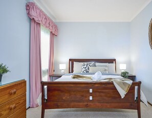 Een net opgemaakt bed met een houten hoofdbord, twee nachtkastjes met lampen, een houten dressoir en een roze gordijn.