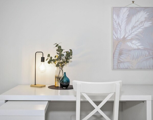 Un bureau blanc avec une lampe au design moderne, un vase avec une plante et une œuvre d’art accrochée au mur.
