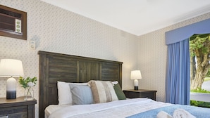Una habitación de hotel con cabecera de madera, dos lámparas de noche, cama con ropa blanca y una ventana con cortinas azules.
