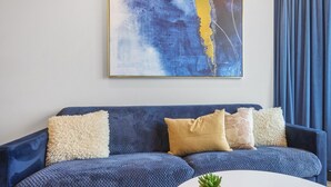 Un sofá azul con tela texturizada, una mesa de centro blanca con un posavasos a cuadros y una planta pequeña, y un cuadro abstracto grande en la pared.