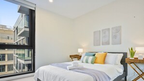Ein modernes Hotelzimmer mit einem großen Bett, Blick auf die Stadt durch ein raumhohes Fenster und einem kleinen Nachttisch mit Lampe.