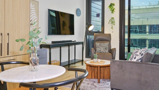 Ein modernes Wohnzimmer mit einem runden Esstisch, einer Couch, einem Fernseher und einem großen Fenster mit Blick auf ein Gebäude.