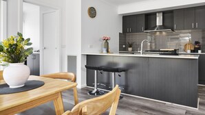 Eine moderne Küche mit dunklen Schränken, einer Edelstahlspüle und einer zentralen Kochinsel. Es gibt einen Essbereich mit einem Holztisch und Stühlen, und eine Wandvase mit Blumen.