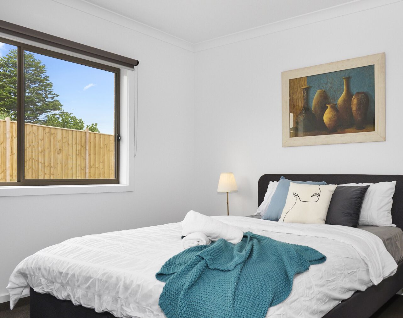A bedroom with a large window, a bed with white bedding, a teal throw blanket, and a painting on the wall.