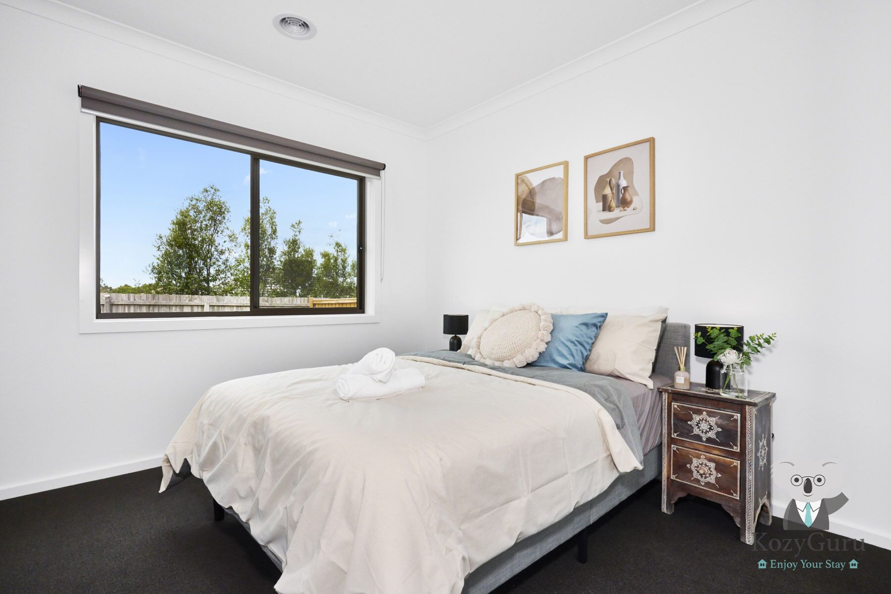 A bedroom with a large window, a bed with white linens, a nightstand with a lamp, and two framed pictures on the wall.