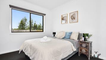 A bedroom with a large window, a bed with white linens, a nightstand with a lamp, and two framed pictures on the wall.
