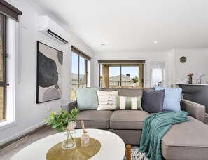A modern living room with a grey sectional sofa, a round coffee table, and a view of the outside. - Kozyguru | Pakenham | Comfortable 4-Bedroom House (Pakenham)