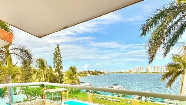A balcony with a view of a swimming pool, palm trees, and a distant city skyline. - Cozy apartment in front of the lagoon in hotel zone (Cancún)