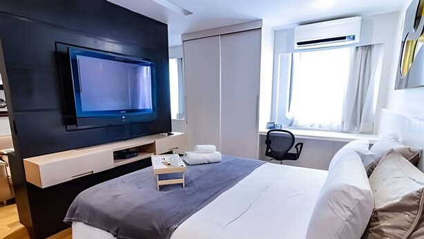 Una habitación de hotel moderna con televisor de pantalla plana, cama con ropa de cama blanca y gris, una mesita de noche con lámpara y ventana con equipo de aire acondicionado.