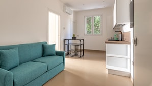 Un salon moderne avec un canapé de couleur turquoise, une petite table noire agrémentée d’une plante, et un coin cuisine doté de meubles blancs et d’une cuisinière.