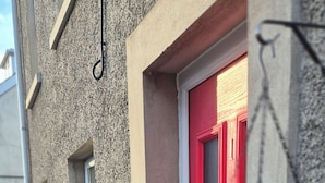 A building exterior with a red door, two potted plants, and a hanging planter with flowers.
