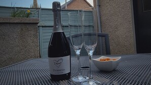 A bottle of Prosecco wine, two wine glasses, and a bowl of snacks on a metal table outdoors.