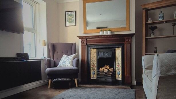 A living room with a fireplace, wooden flooring, a grey rug, a grey armchair, and a sofa.